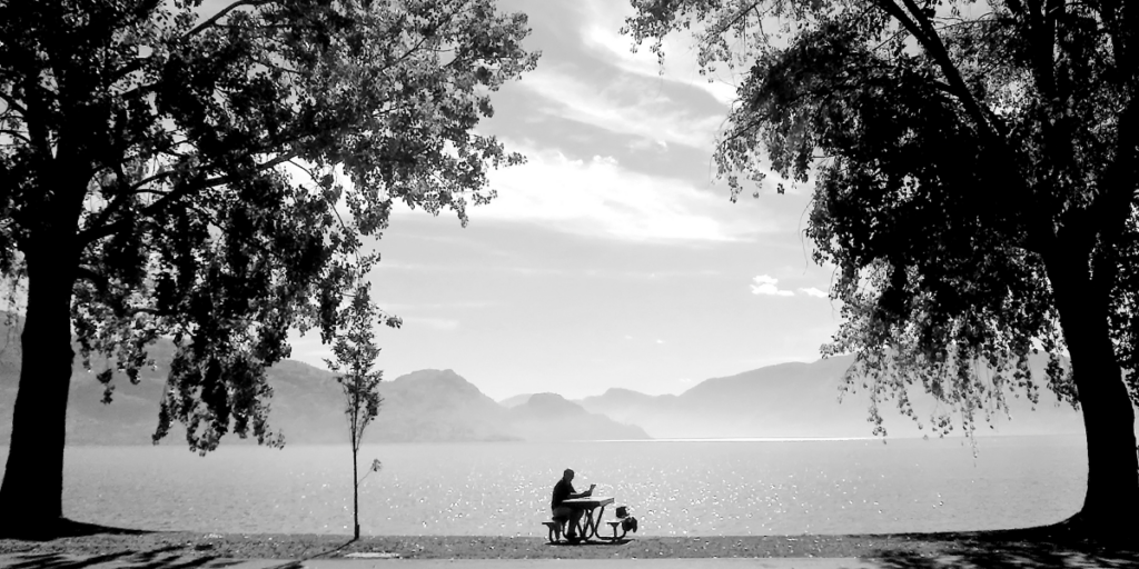 A person reading and sitting at a picnic bench with large trees, a lake, and mountains in the background, in black and white.