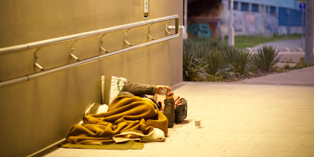 Homeless person laying on their side underneath a railing covered with a blanket and using their phone.