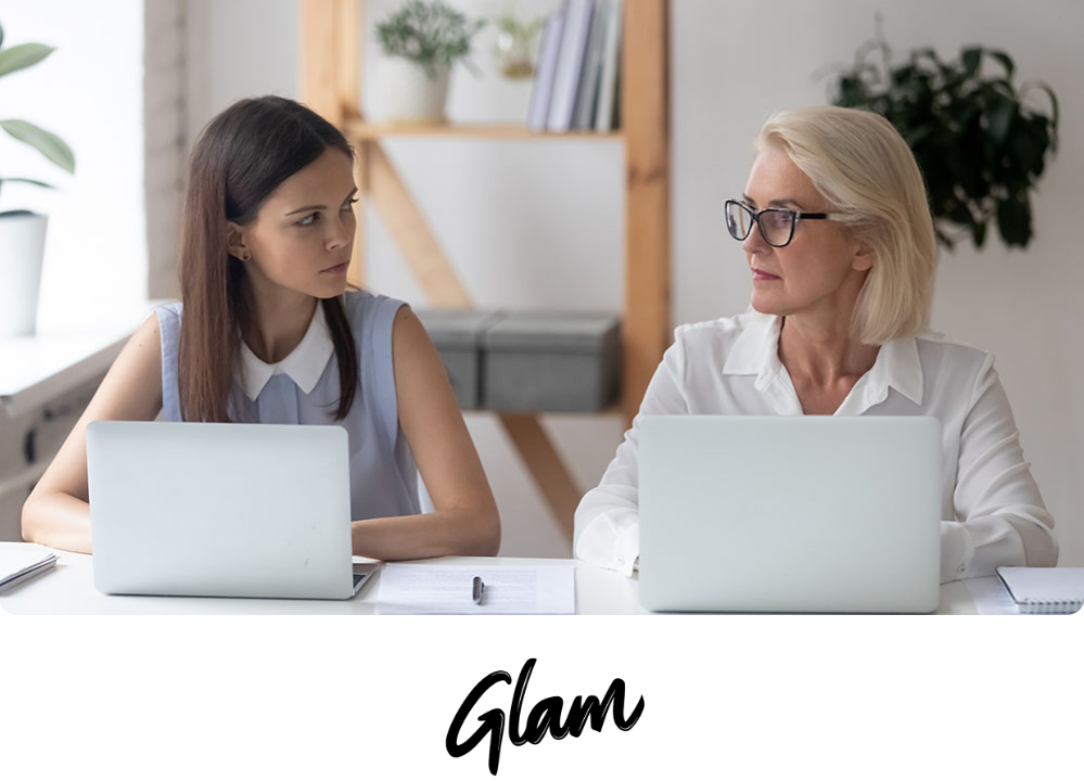 Stock image of two businesswomen looking at each other as they sit in front of their computers.