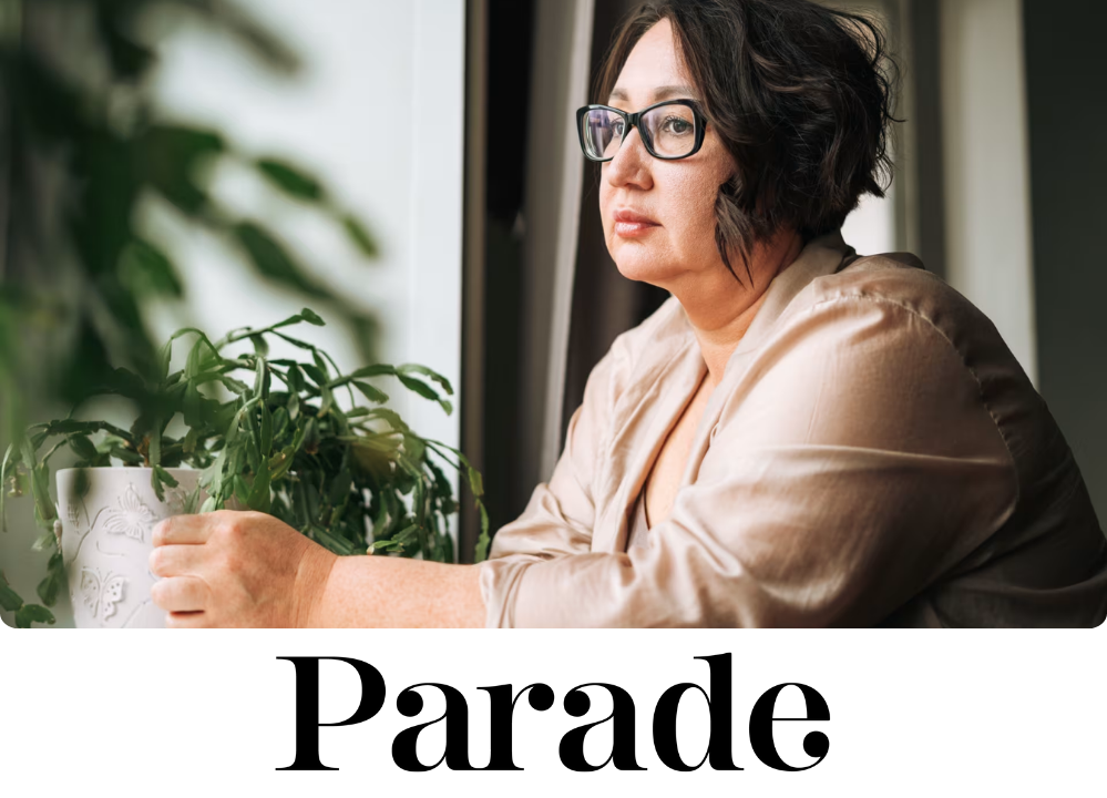 Stock image of a woman with short dark hair and glasses looking thoughtfully out of a window while resting her hands near a potted plant.