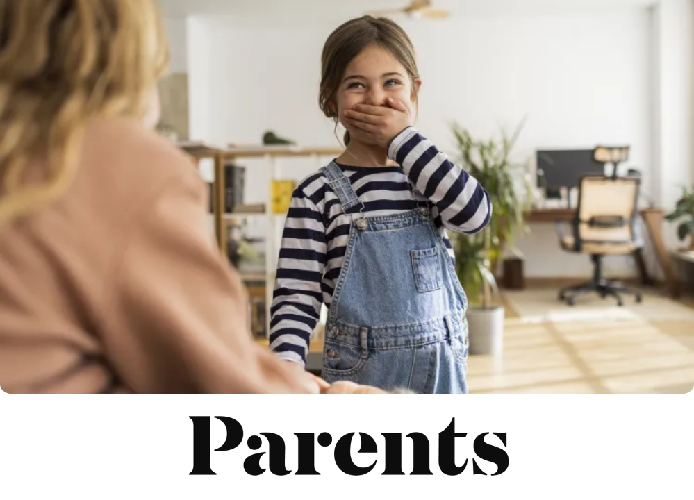 Stock image of a young girl in denim overalls covers her mouth while giggling at an adult sitting in front of her.
