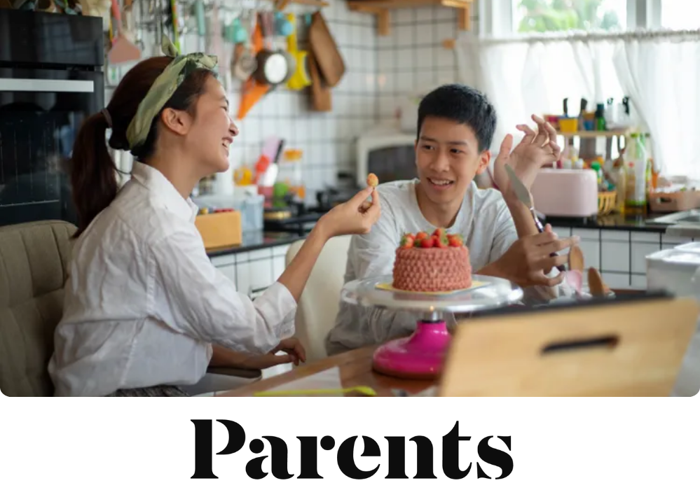 Stock image of a girl laughing while holding a strawberry toward a boy as they sit together in a kitchen with a pink cake.
