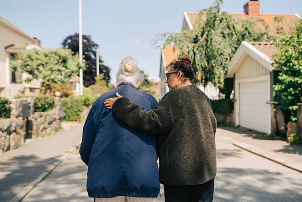 https-media-gettyimages-com-id-2224134490-photo-rear-view-of-female-caregiver-assisting-senior-woman-walking-on-street-at-sunny-day